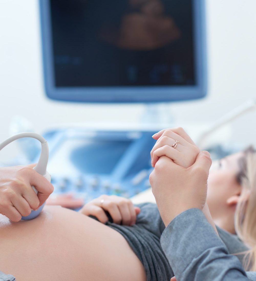 Female doctor touching stomach by tool of woman who lying,showing baby at screen of computer. Professional doctor using ultrasound equipment screening of pregnant woman. Husband holding hand his wife.